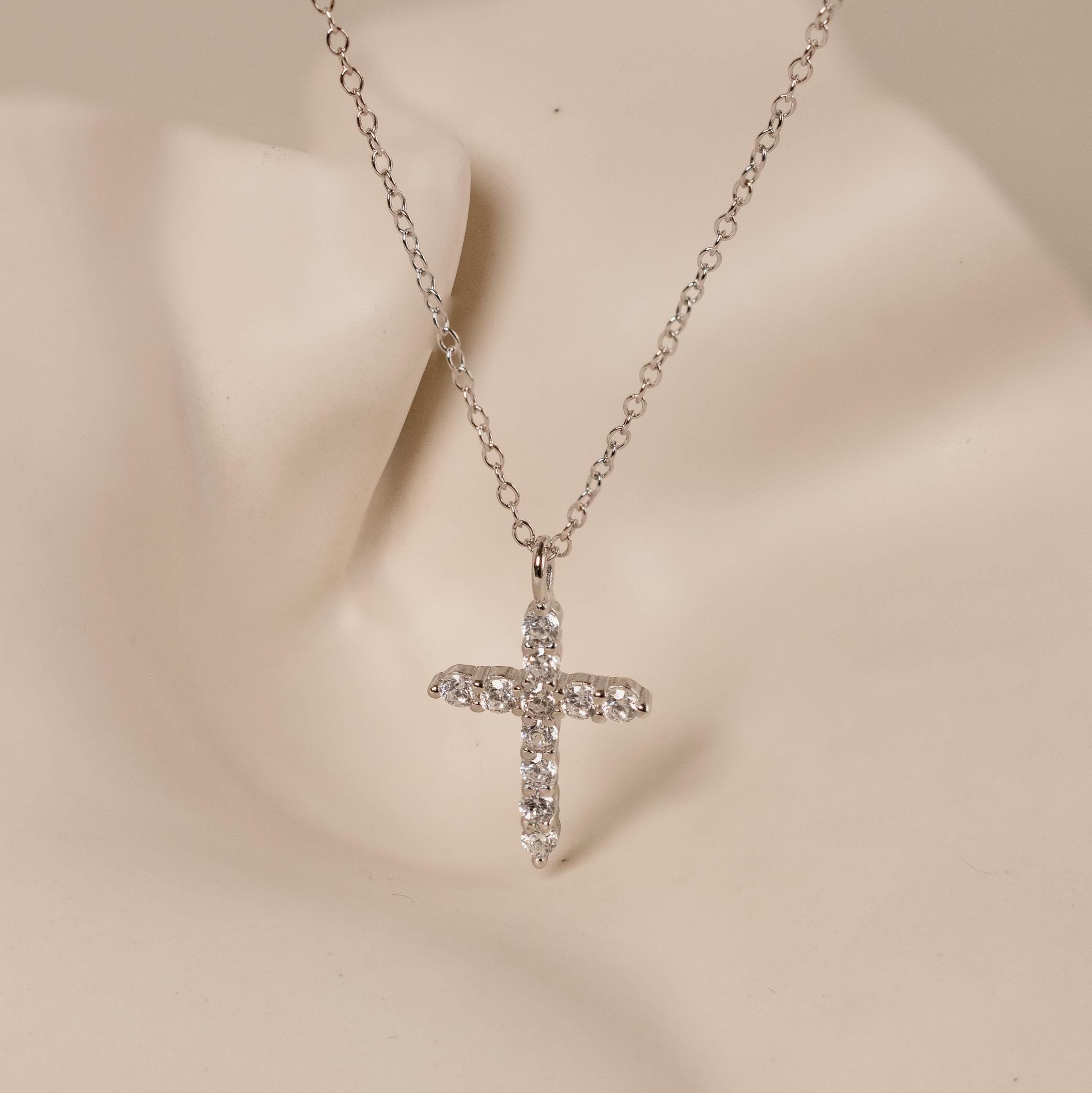 The Pave Cross Necklace features a silver chain and rhinestone cross pendant, elegantly displayed on a neutral mannequin bust.