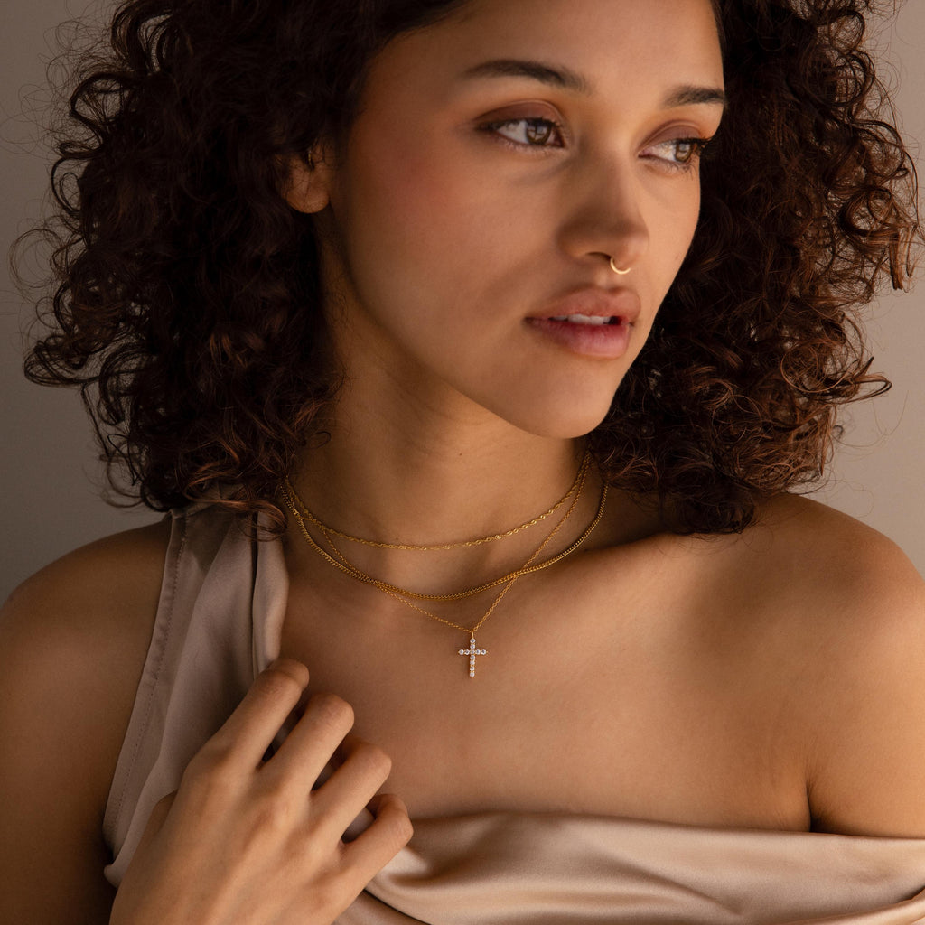 A woman with curly hair gazes thoughtfully to the side, wearing layered gold necklaces featuring the Pave Cross Necklace—an elegant and exquisite jewelry look.