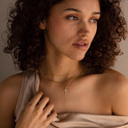 A woman with curly hair gazes thoughtfully to the side, wearing layered gold necklaces featuring the Pave Cross Necklace—an elegant and exquisite jewelry look.