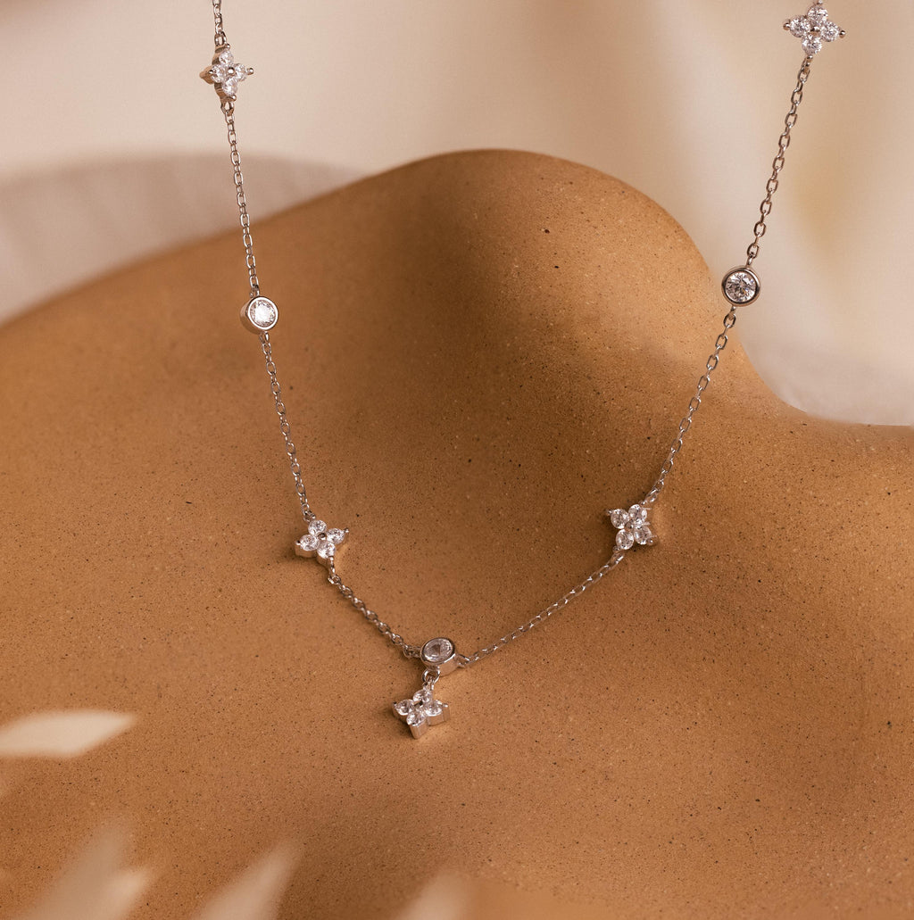 The Ivy Diamond Station Necklace, featuring delicate silver with small star and round pendants accented by diamonds, is shown draped on a smooth tan surface.
