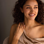 A smiling woman in a draped beige top poses with her hand near her collarbone, wearing layered gold necklaces featuring the Ivy Diamond Station Necklace.