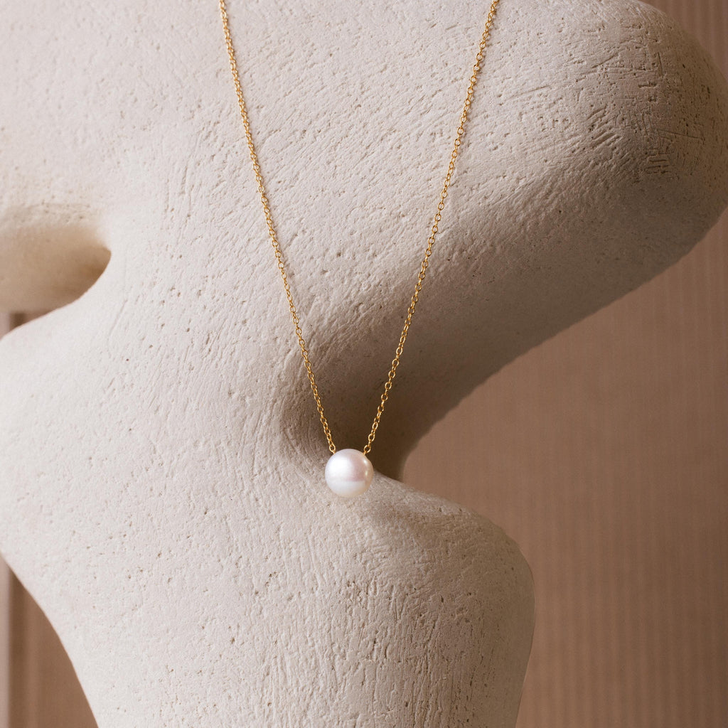 The Classic Pearl Pendant Necklace features a single white pearl on a dainty gold chain, displayed against a textured beige sculpture. This minimalist piece adds an elegant touch to any look.