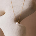 The Classic Pearl Pendant Necklace features a single white pearl on a dainty gold chain, displayed against a textured beige sculpture. This minimalist piece adds an elegant touch to any look.