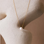 The Classic Pearl Pendant Necklace features a single white pearl on a dainty gold chain, displayed against a textured beige sculpture. This minimalist piece adds an elegant touch to any look.