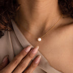 A woman wearing the Classic Pearl Pendant Necklace, which features a delicate chain and a single pearl pendant, gently touches her collarbone.