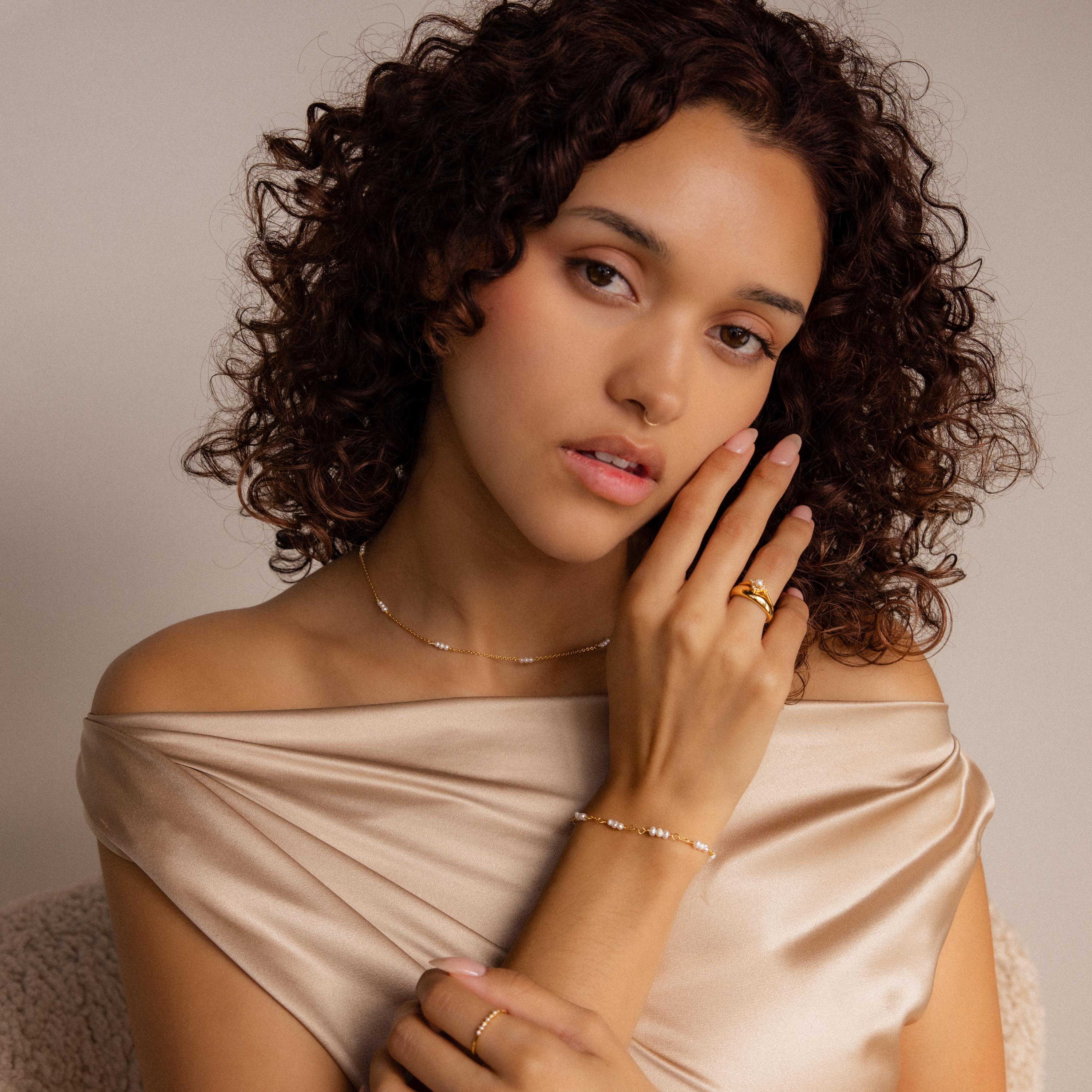 A woman with curly hair wears a satin off-shoulder top and gold jewelry, featuring the Hali Pearl Station Necklace, as she poses thoughtfully.