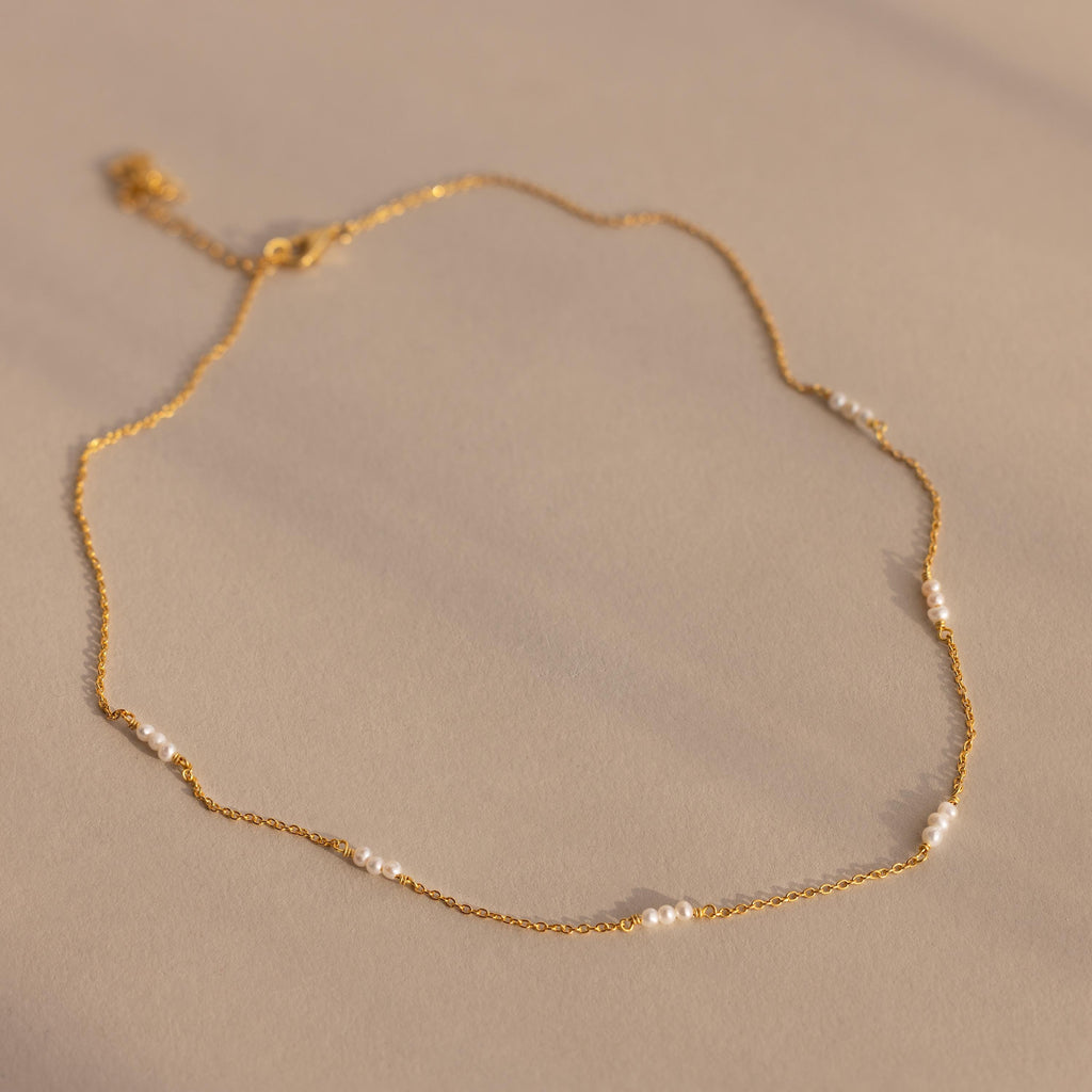 The Hali Pearl Station Necklace features delicate gold links with small white pearls, shown on a neutral beige background—ideal for those who love minimalist jewelry.