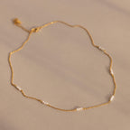 The Hali Pearl Station Necklace features delicate gold links with small white pearls, shown on a neutral beige background—ideal for those who love minimalist jewelry.