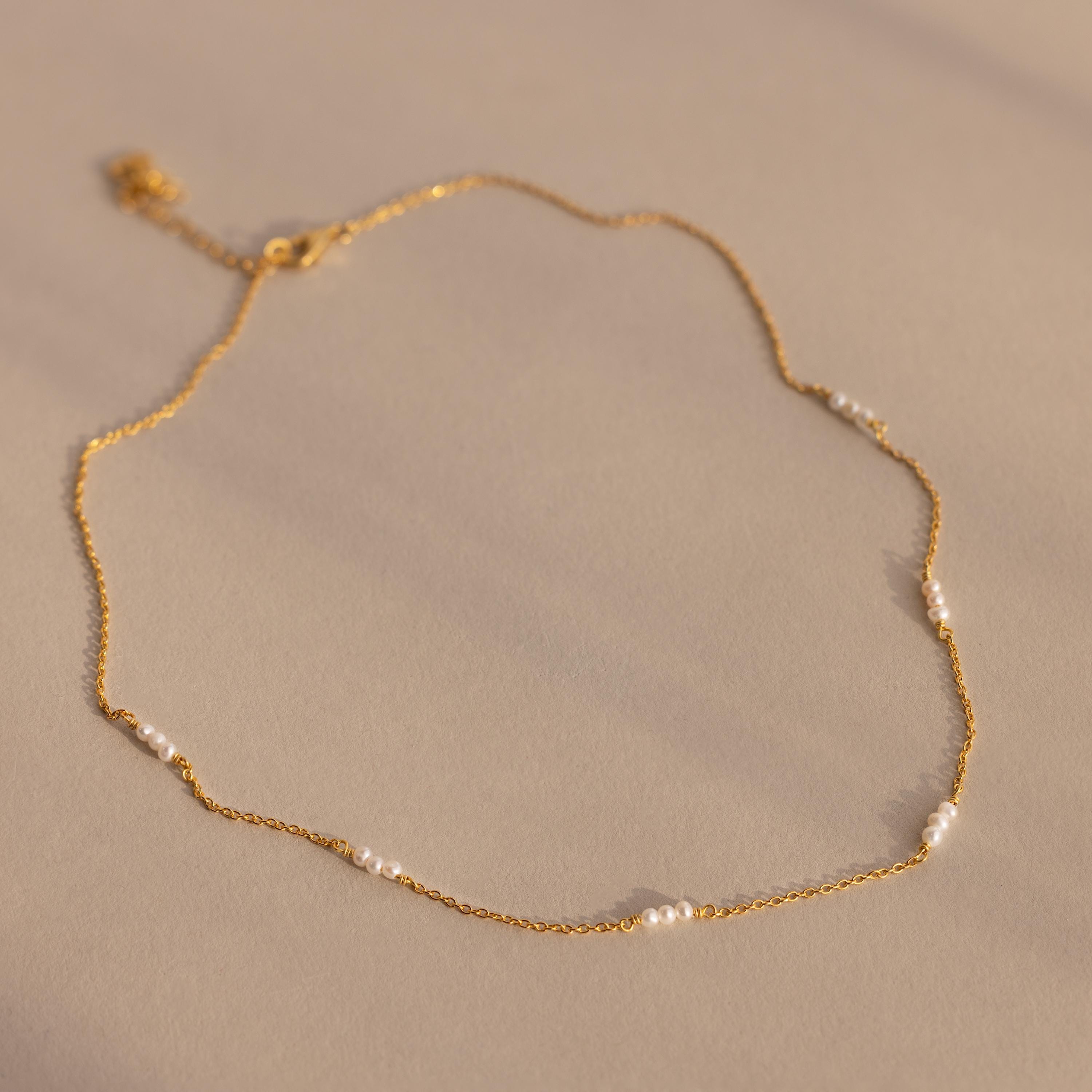 The Hali Pearl Station Necklace features delicate gold links with small white pearls, shown on a neutral beige background—ideal for those who love minimalist jewelry.