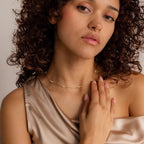 Woman with curly hair wearing the Hali Pearl Station Necklace and a satin top, touching her chest while looking at the camera.