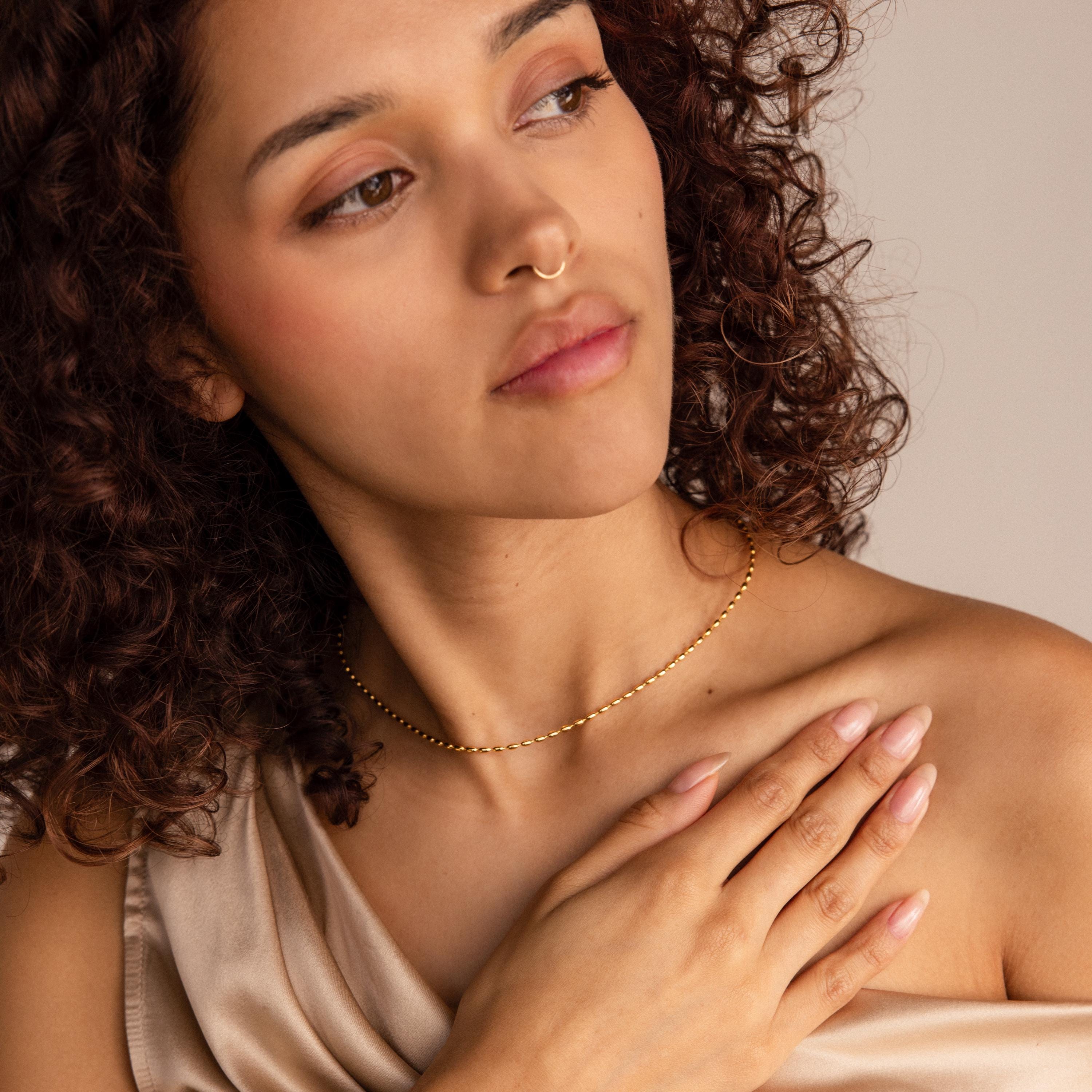 A woman with curly hair, wearing an off-shoulder top and the Oval Beaded Necklace, touches her chest while gazing to the side.