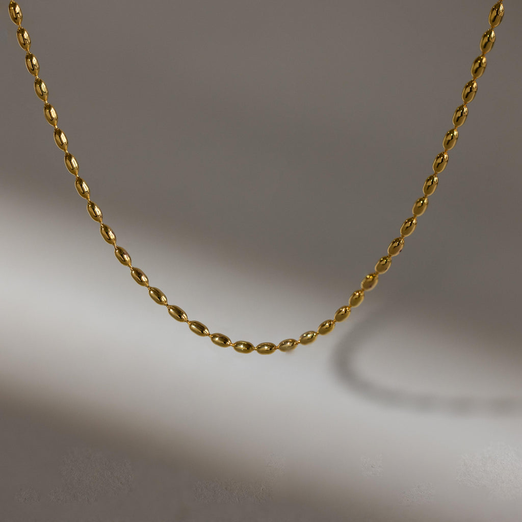 The Oval Beaded Necklace features a simple gold design with a twisted chain, displayed against a gray background and casting a soft shadow.