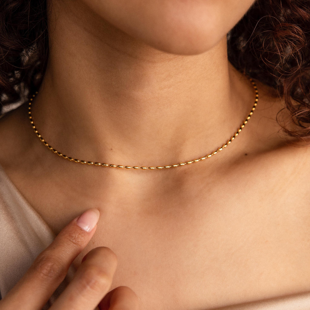 A woman wearing the Oval Beaded Necklace—a delicate gold chain—gently touches her collarbone with her finger.