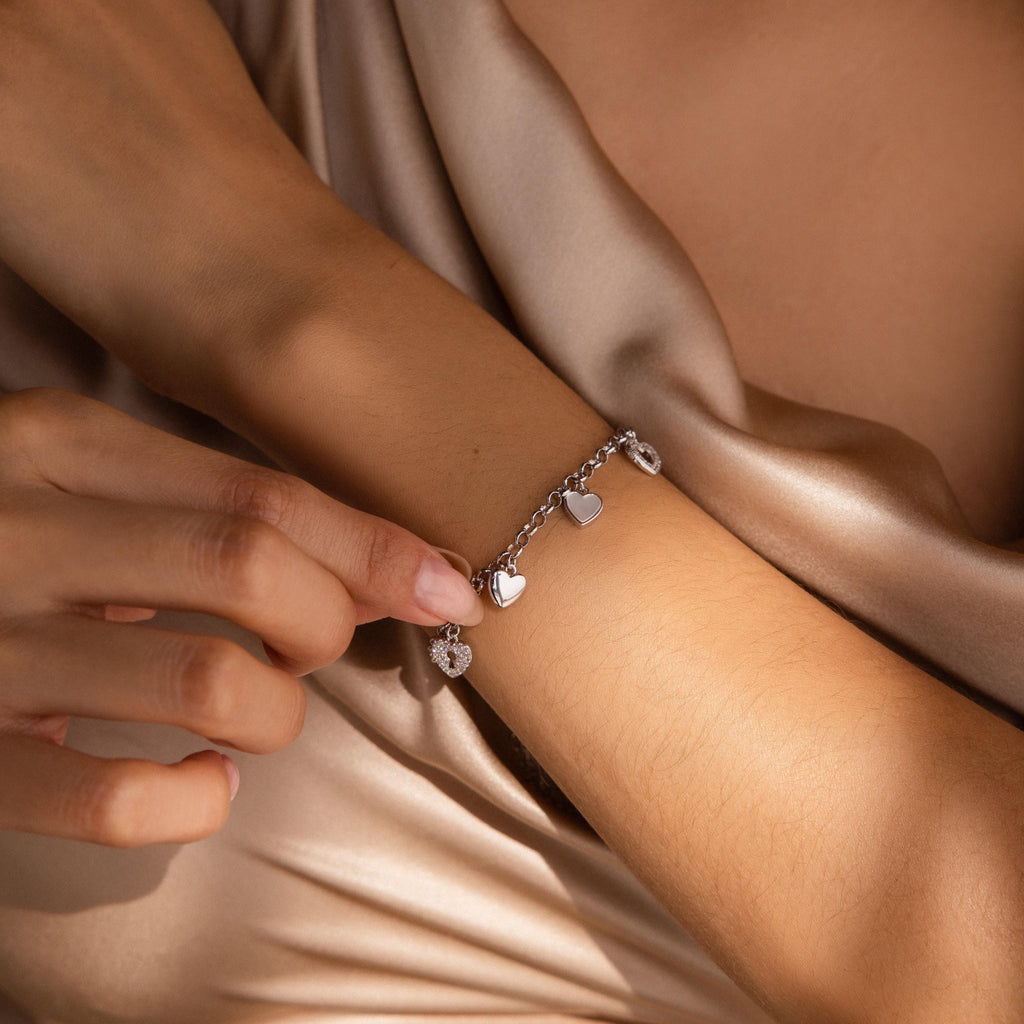 A hand touches the Heart Charm Bracelet, featuring delicate heart charms and worn on a wrist resting on silk—an ideal gift for your girlfriend.