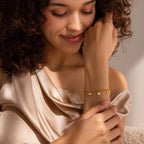 A woman in a satin top smiles, admiring the Heart Charm Bracelet in 18K Gold as its heart charms catch the light—an ideal gift for your girlfriend.