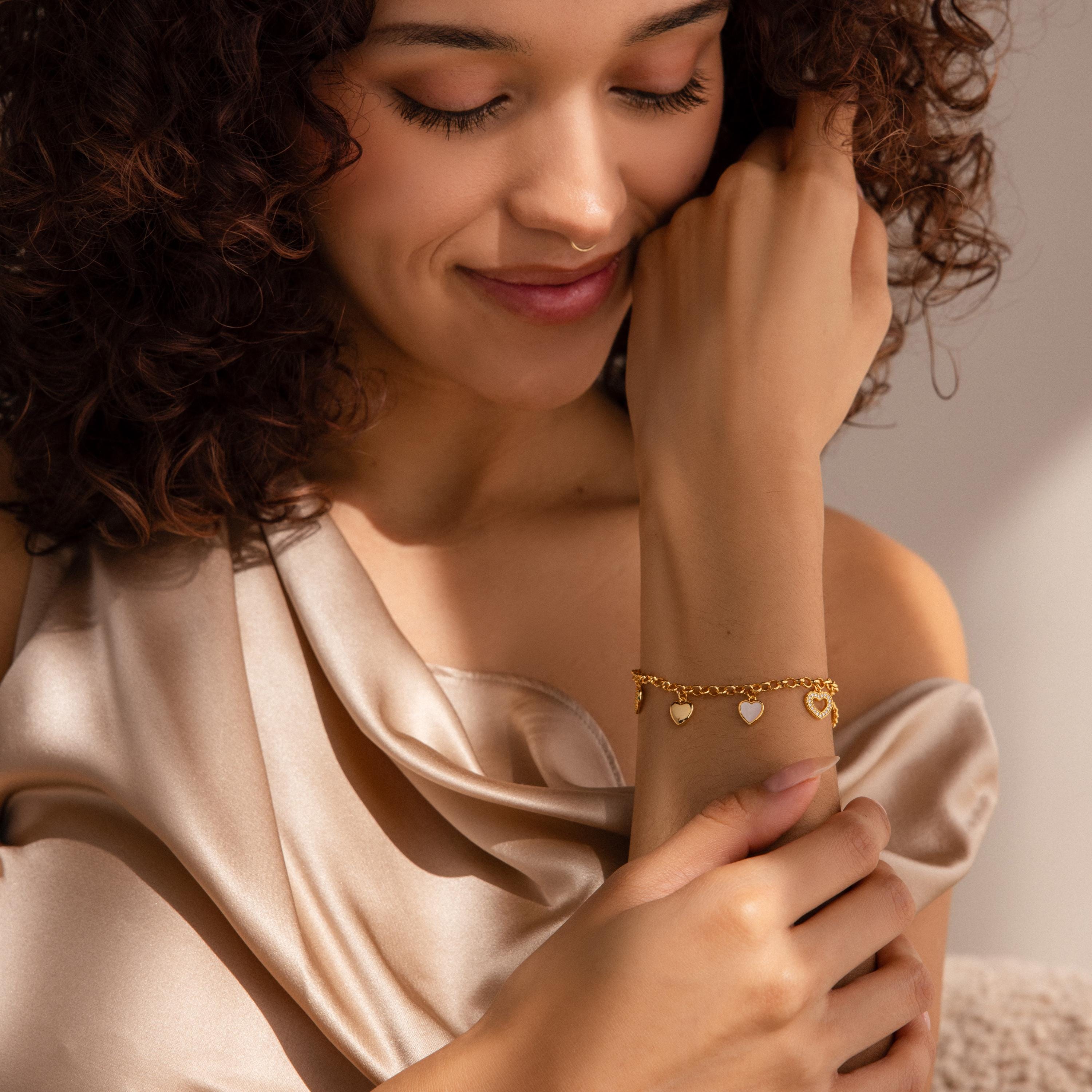 A woman in a satin top smiles, admiring the Heart Charm Bracelet in 18K Gold as its heart charms catch the light—an ideal gift for your girlfriend.