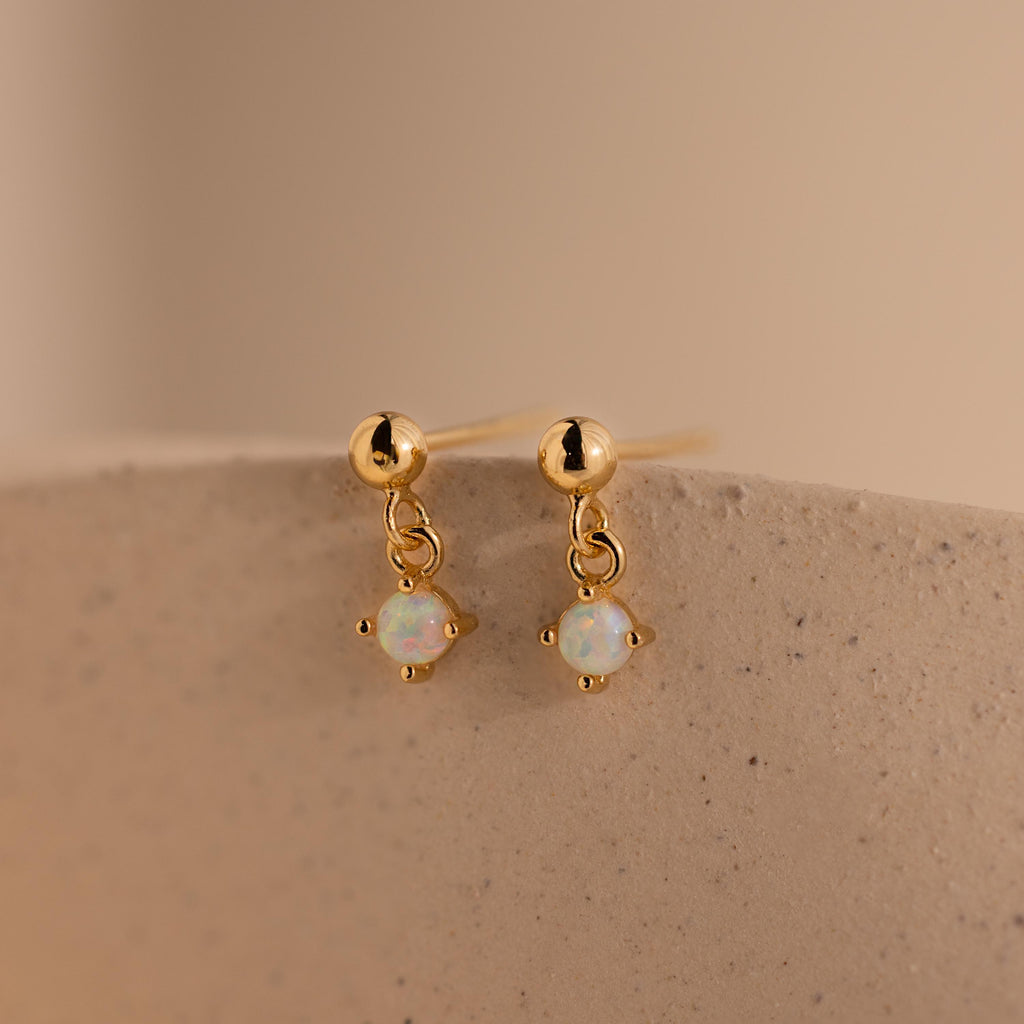 Dainty Opal Drop Studs showcase small round opal gemstone dangles on gold, displayed on a textured beige surface. Lightweight and elegant, these earrings are perfect for effortless style.