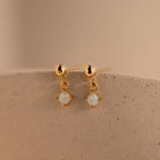 Dainty Opal Drop Studs showcase small round opal gemstone dangles on gold, displayed on a textured beige surface. Lightweight and elegant, these earrings are perfect for effortless style.