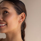 A woman with brown hair in a ponytail smiles, wearing Dainty Opal Drop Studs that add a lightweight elegance to her look.
