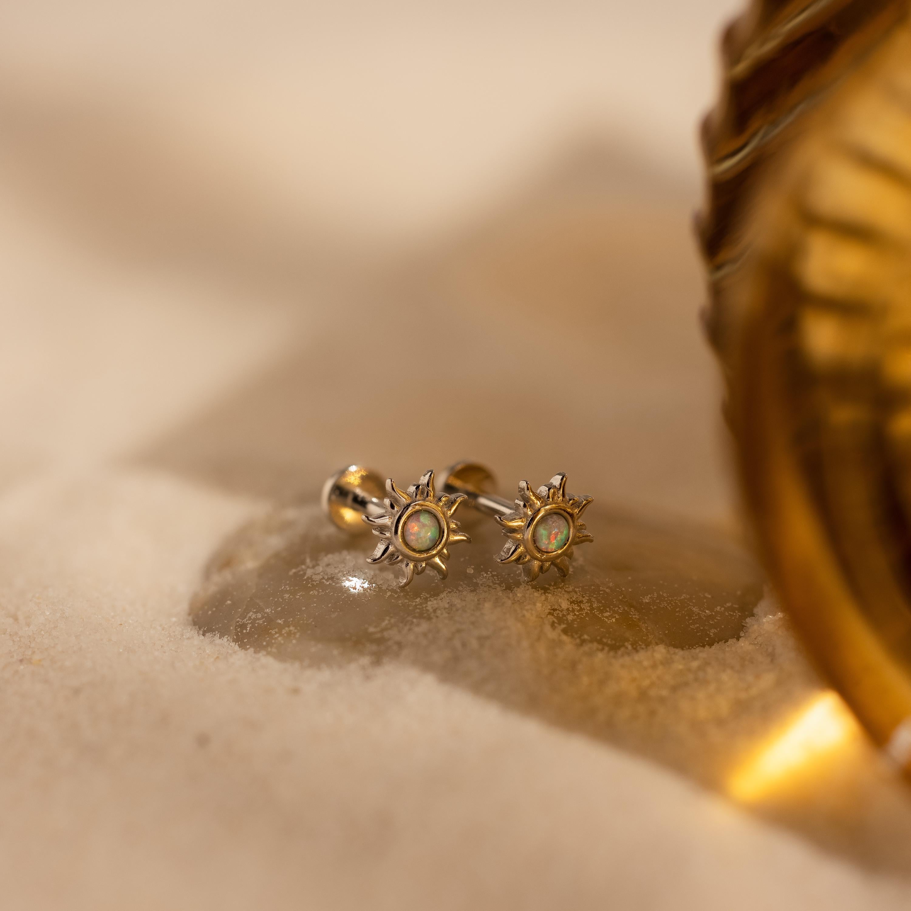 Celestial jewelry at its finest—the Opal Sun Flat Back Studs, featuring vibrant green stones and a flat back design, elegantly rest on sand beside a golden decorative object.