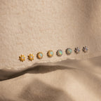 Eight pairs of Pave Opal Moon Studs in gold and silver shine with celestial sparkle on a textured beige surface.