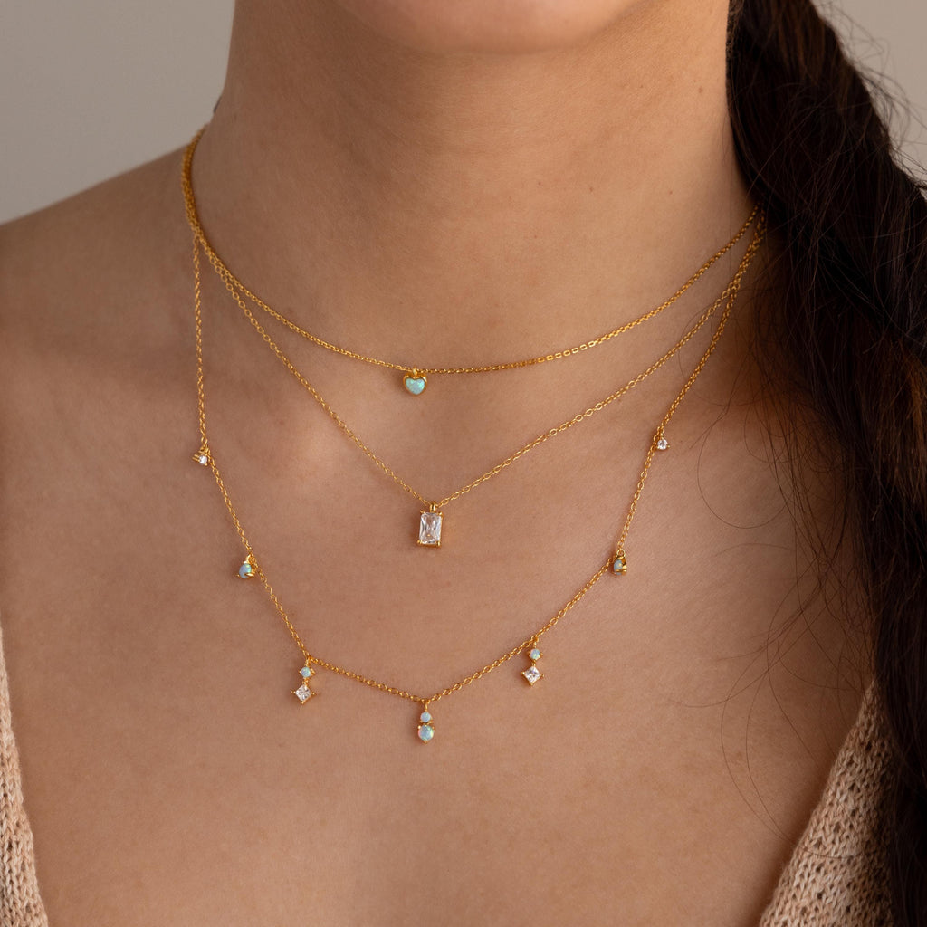 The Opal Diamond Station Necklace shines on her neck, its three gold layers with opal and gemstone pendants perfect for bridesmaids or a birthstone gift. Worn with a beige top, it highlights the necklace's elegance.