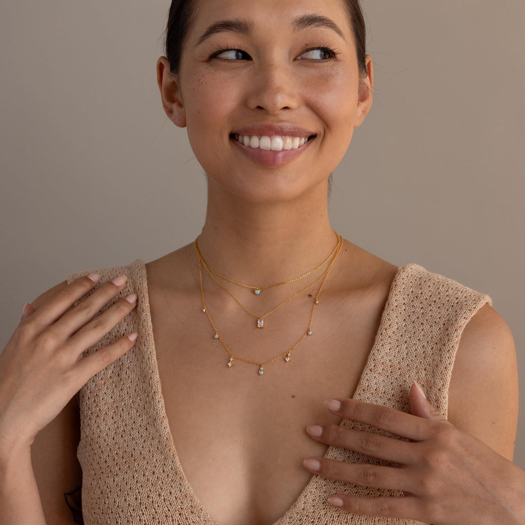 Smiling woman wearing layered gold necklaces, including the Opal Diamond Station Necklace, with a sleeveless beige knit top against a neutral background.