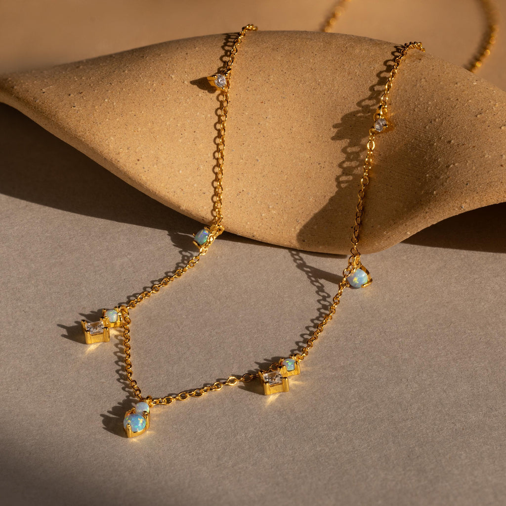 The Opal Diamond Station Necklace features blue opal charms on a gold chain, making it an elegant birthstone gift or bridesmaid keepsake, shown on a textured beige surface in sunlight.