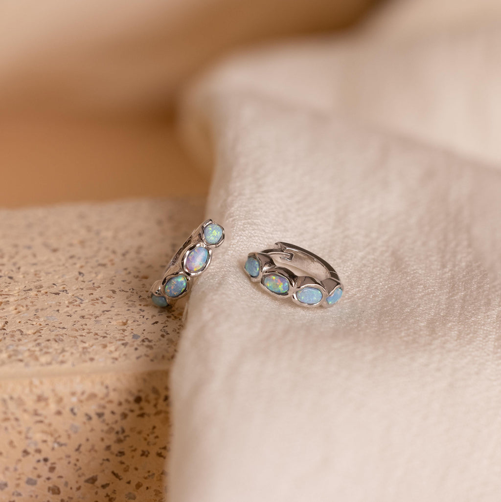 Blue Opal Beaded Huggies featuring iridescent blue opal stones on soft cream-colored fabric—these earrings make stunning birthstone gifts for any occasion.