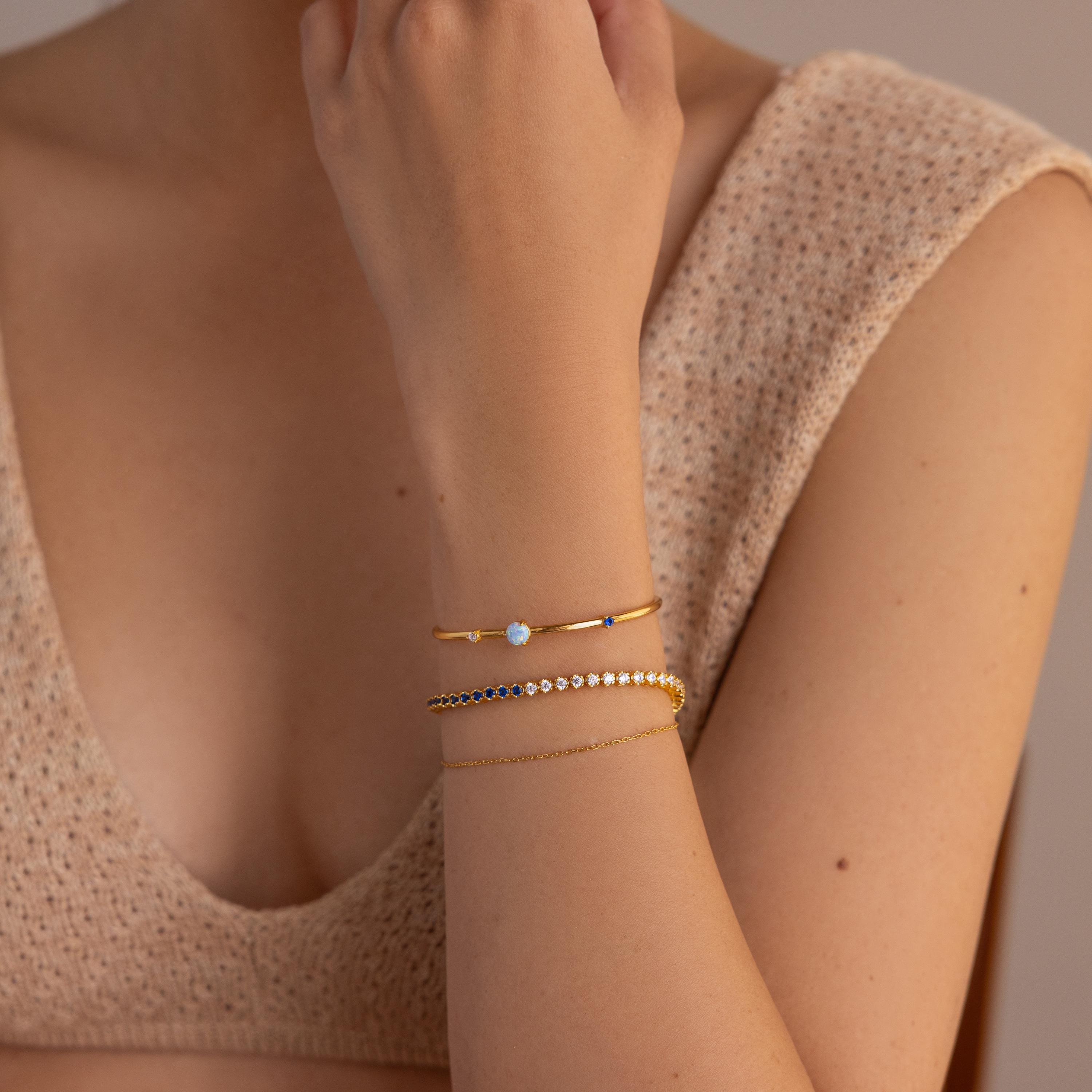 A woman wears a beige knit top and three delicate gold bracelets on her wrist, including the Opal & Diamond Cuff Bracelet featuring a small opal stone.
