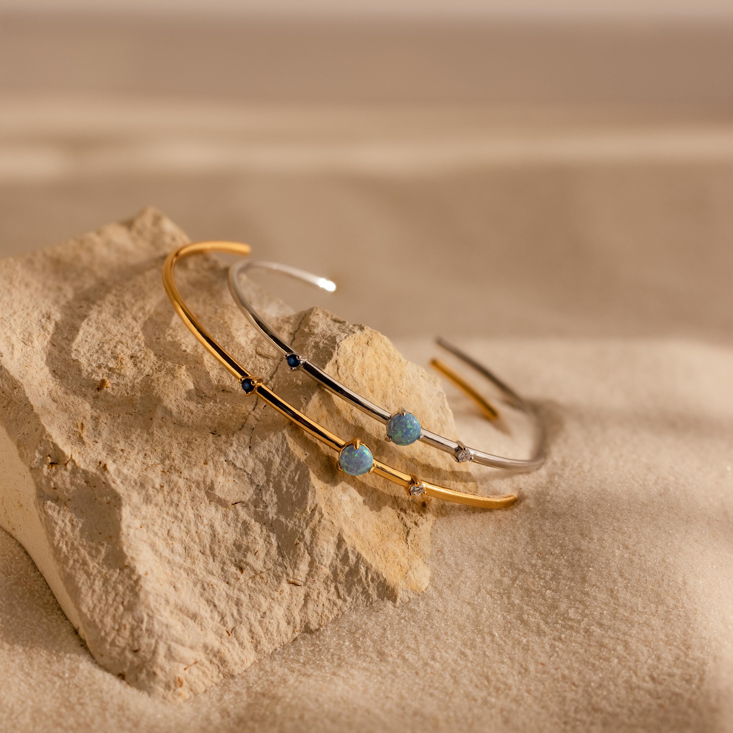 An Opal & Diamond Cuff Bracelet rests on a textured rock, its gemstones glimmering against a sandy background.