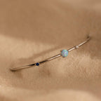 The Opal & Diamond Cuff Bracelet, featuring blue, opal, and clear gemstones, rests delicately on soft beige sand.