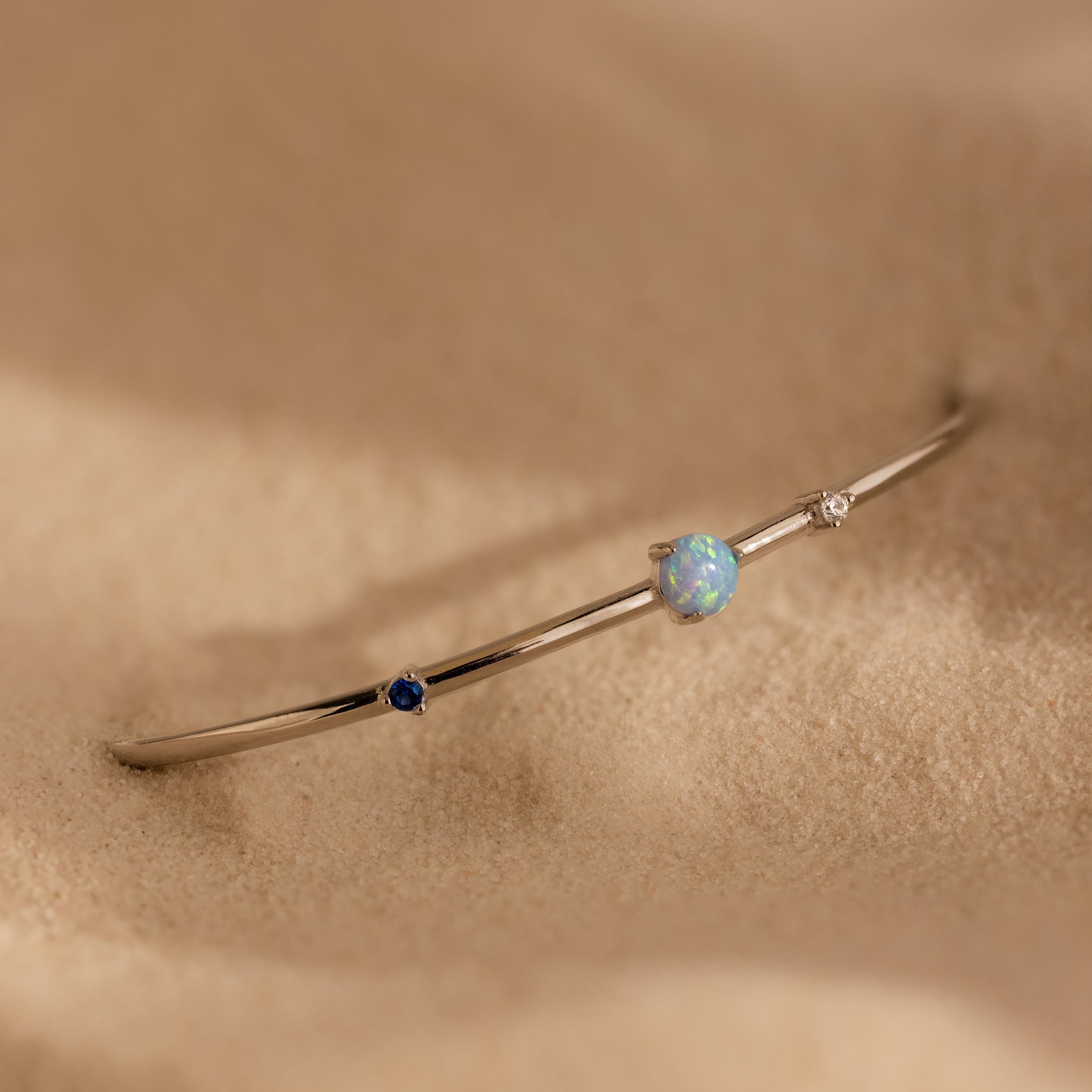 The Opal & Diamond Cuff Bracelet, featuring blue, opal, and clear gemstones, rests delicately on soft beige sand.