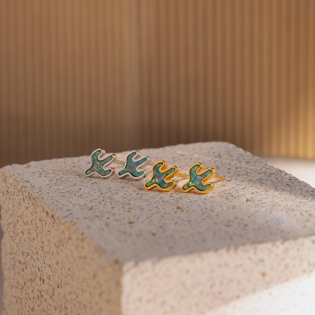 Four Opal Cactus Studs, each featuring a green gemstone and bird-inspired design, rest on a textured beige block against a softly lit, striped backdrop.