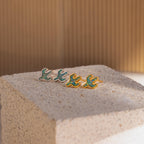 Four Opal Cactus Studs, each featuring a green gemstone and bird-inspired design, rest on a textured beige block against a softly lit, striped backdrop.