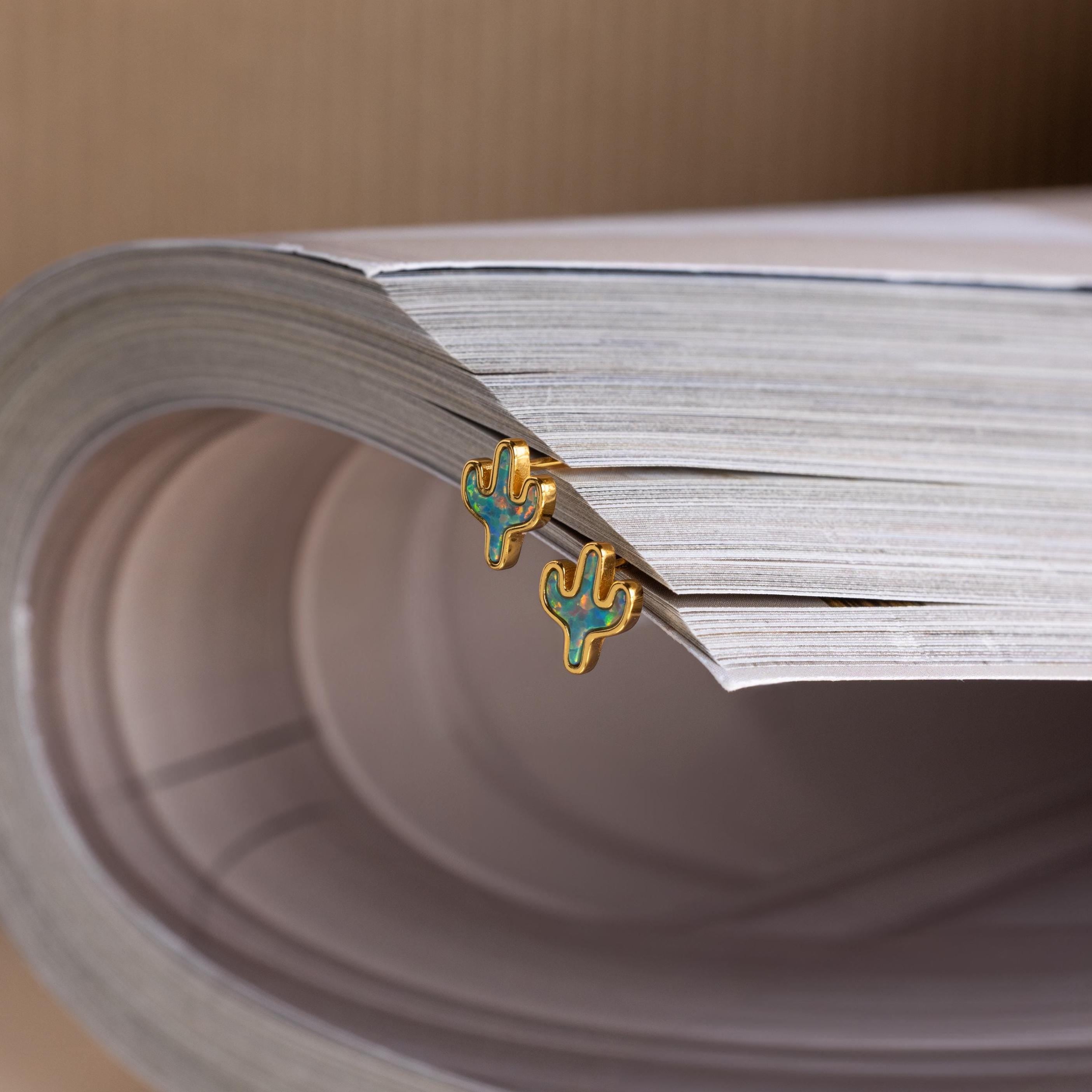 Opal Cactus Studs shimmer in gold and green as they rest delicately atop a stack of white papers.