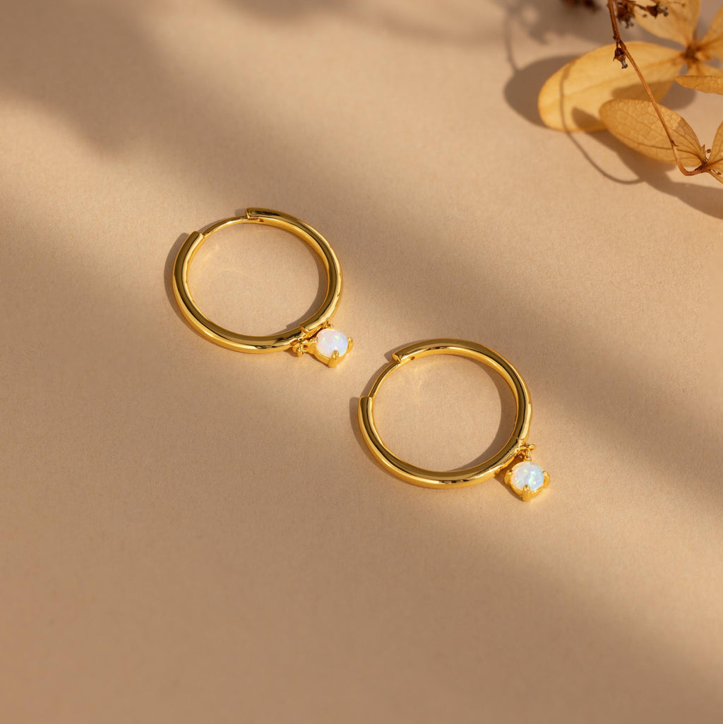 The Dainty Opal Drop Huggies feature gold hoops with small opal-like charms, displayed on a beige surface with soft shadows, making them the perfect pair of delicate earrings.