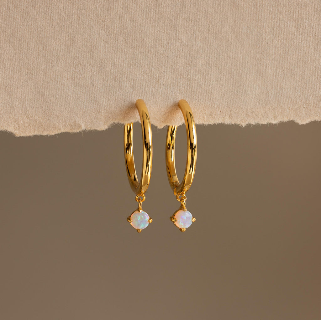 The Dainty Opal Drop Huggies feature gold hoops with small round opal charms and rest on a textured beige surface, creating delicate earrings that add a touch of elegance to your everyday style.