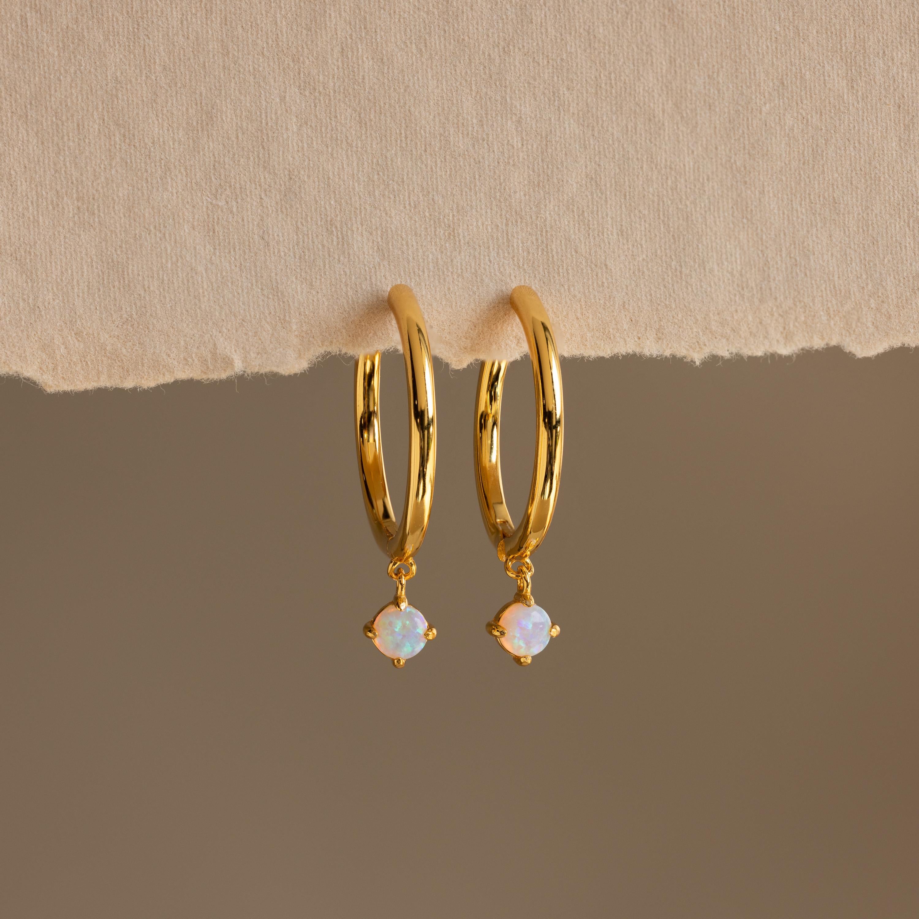 The Dainty Opal Drop Huggies feature gold hoops with small round opal charms and rest on a textured beige surface, creating delicate earrings that add a touch of elegance to your everyday style.