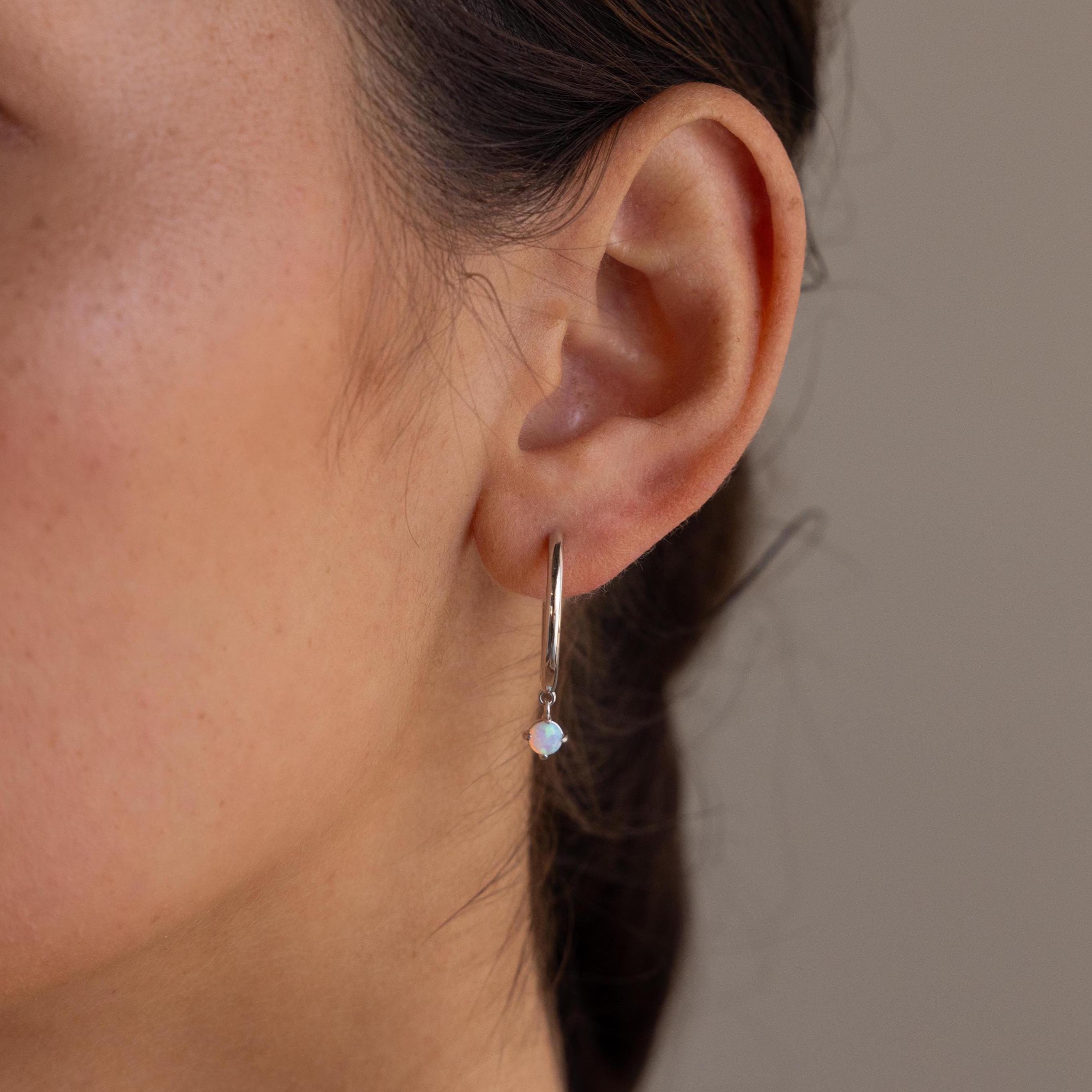 Close-up of a person’s ear wearing Dainty Opal Drop Huggies—small silver hoops adorned with round, light opal stones.
