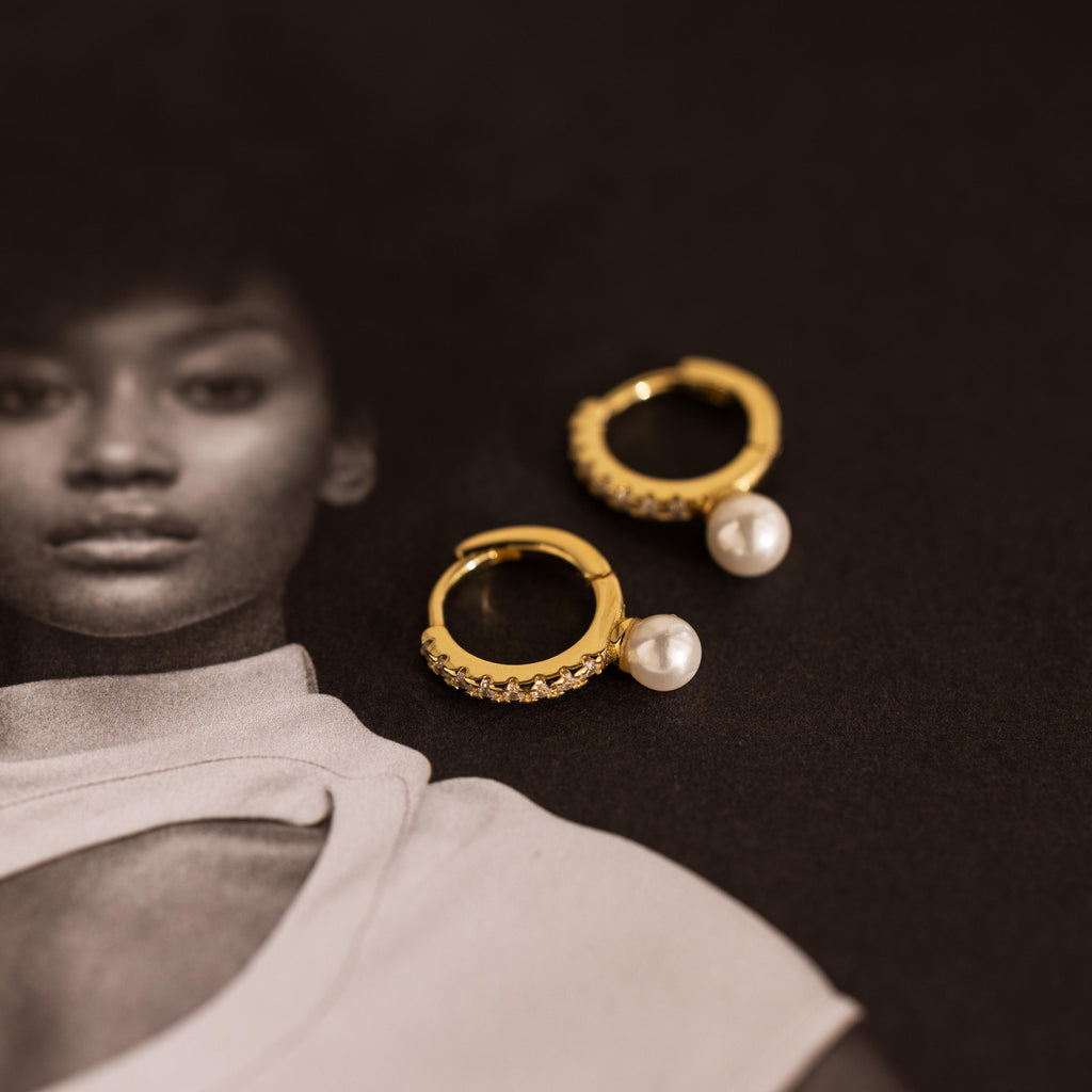 The Pave Pearl Drop Huggies, featuring gold hoops with pearls and crystals, are displayed on a black-and-white photo of a woman in a white top.