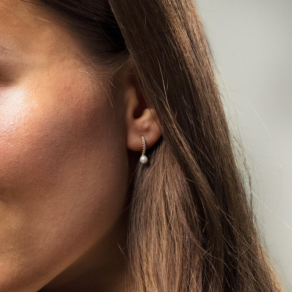 A close-up of a woman's ear adorned with Pave Pearl Drop Huggies in a gold setting, paired with straight brown hair.
