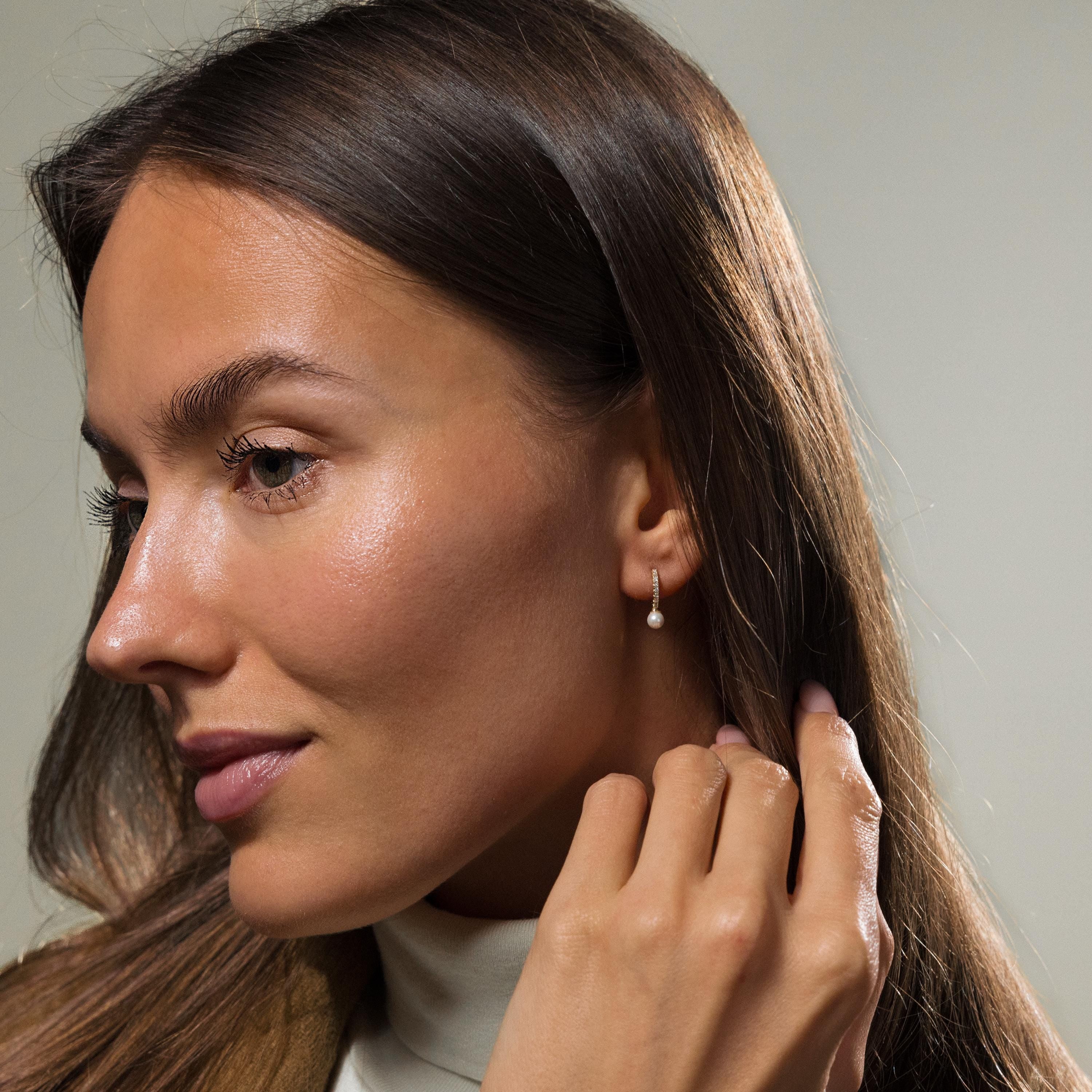 A woman with long brown hair touches her ear and gazes to the side, showcasing the elegant Pave Pearl Drop Huggies.