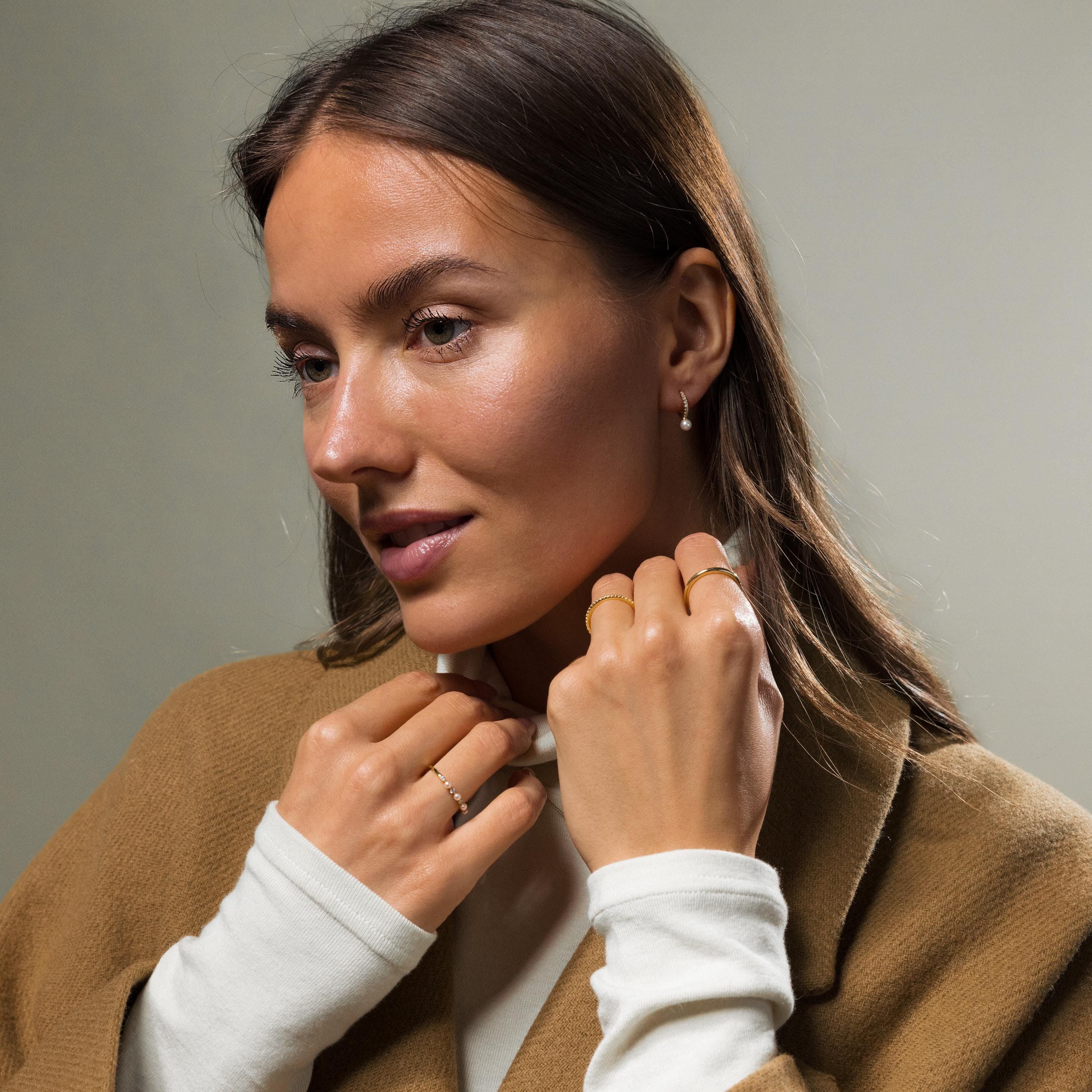 Woman with long brown hair wearing rings, a tan coat over a white top, and the Pave Pearl Drop Huggies, looking to the side.