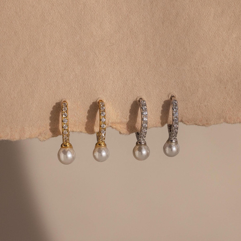 Two pairs of pearl drop earrings, including the Pave Pearl Drop Huggies and a gold pair with pave crystal accents, are displayed on a textured beige background.