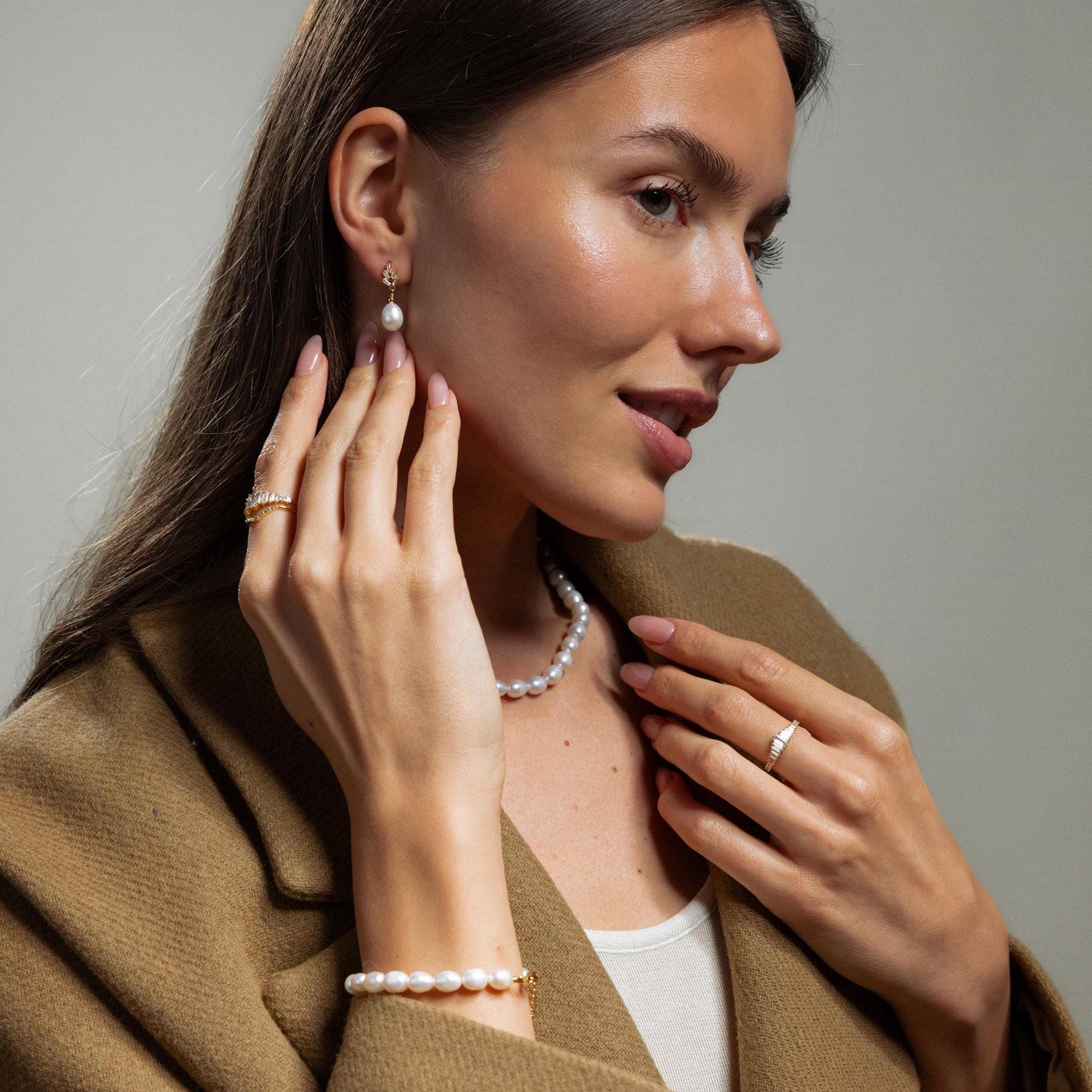 Woman wearing Pearl Diamond Drop Earrings and a brown coat, touching her earring and looking to the side.