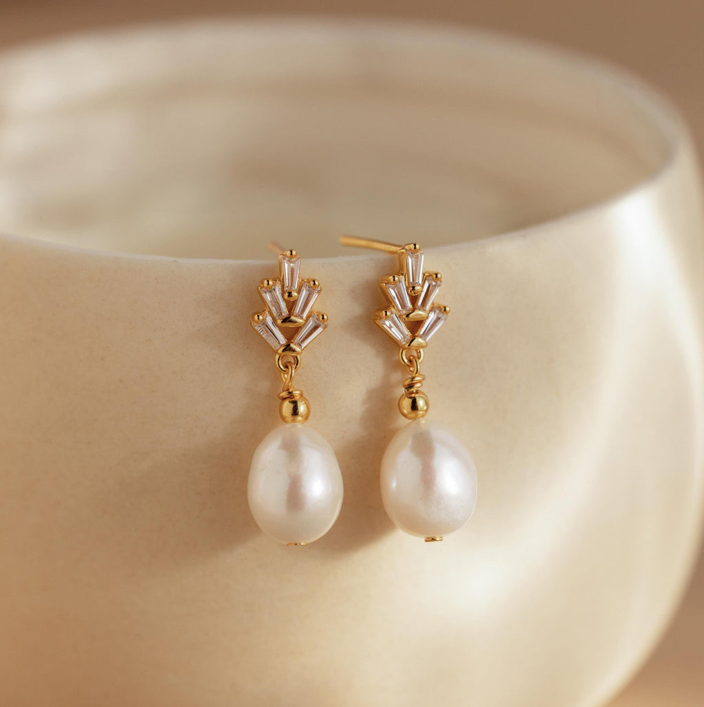 Gold and pearl diamond drop earrings rest on a smooth, cream ceramic surface, highlighting the classic elegance of Pearl Diamond Drop Earrings.