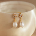 Gold and pearl diamond drop earrings rest on a smooth, cream ceramic surface, highlighting the classic elegance of Pearl Diamond Drop Earrings.