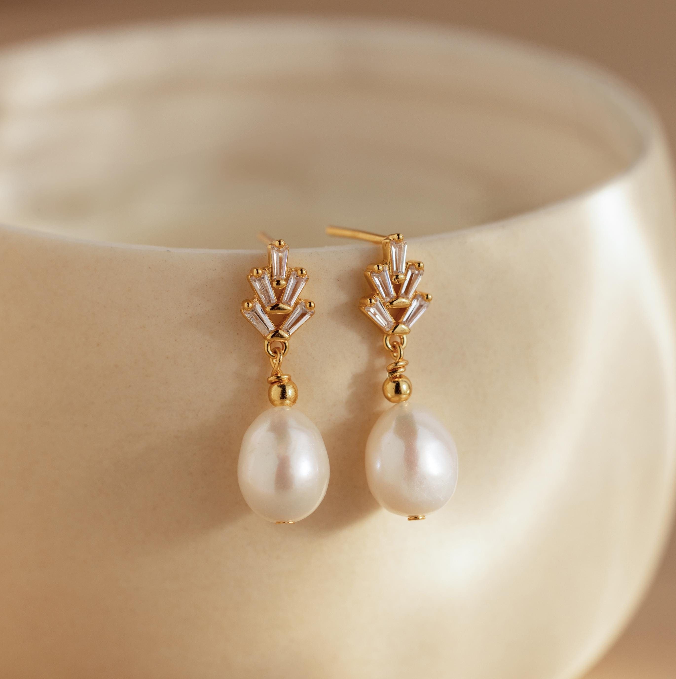 Gold and pearl diamond drop earrings rest on a smooth, cream ceramic surface, highlighting the classic elegance of Pearl Diamond Drop Earrings.