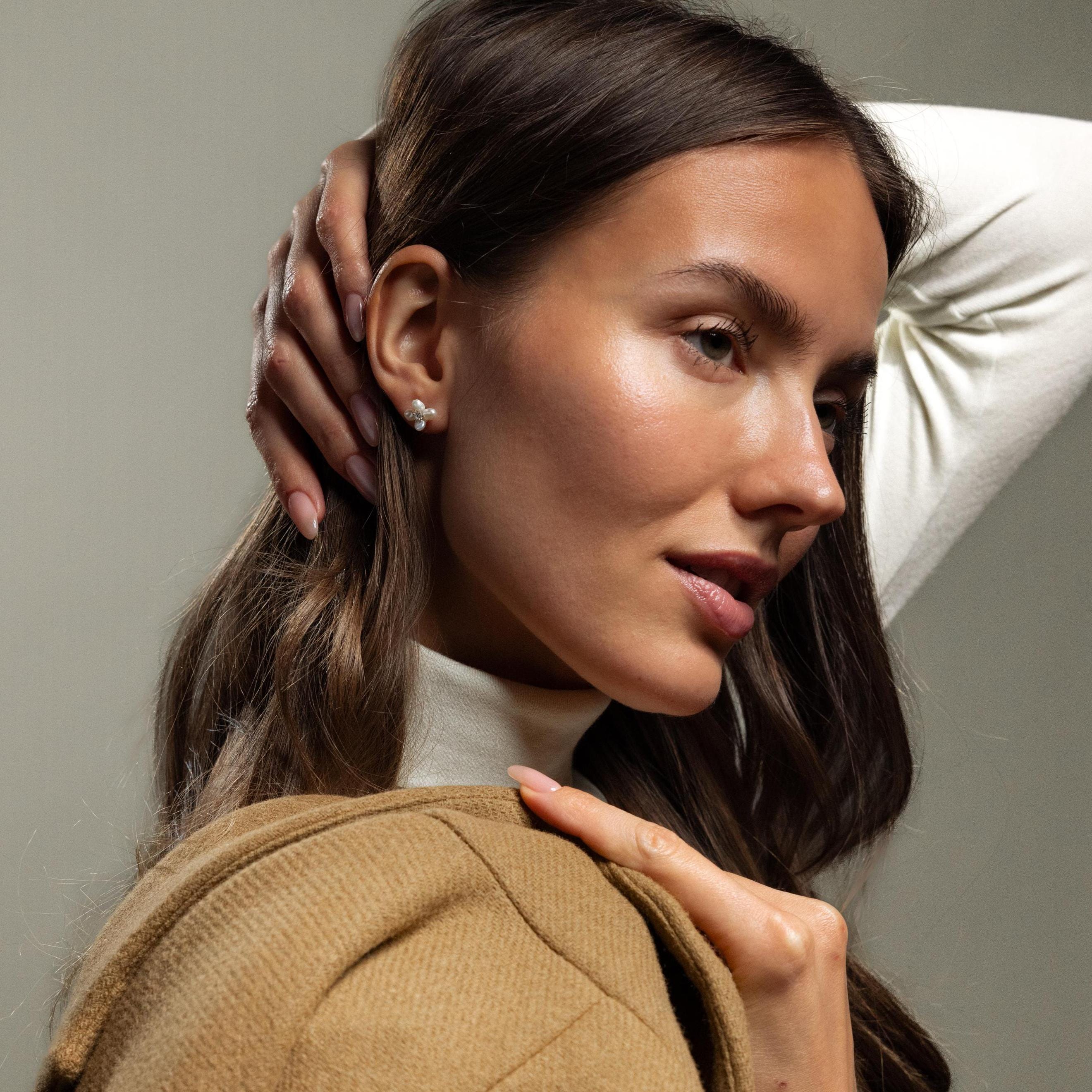 A woman in a white turtleneck and tan coat poses with her hand on her head, highlighting the elegant Pearl Flower Studs.